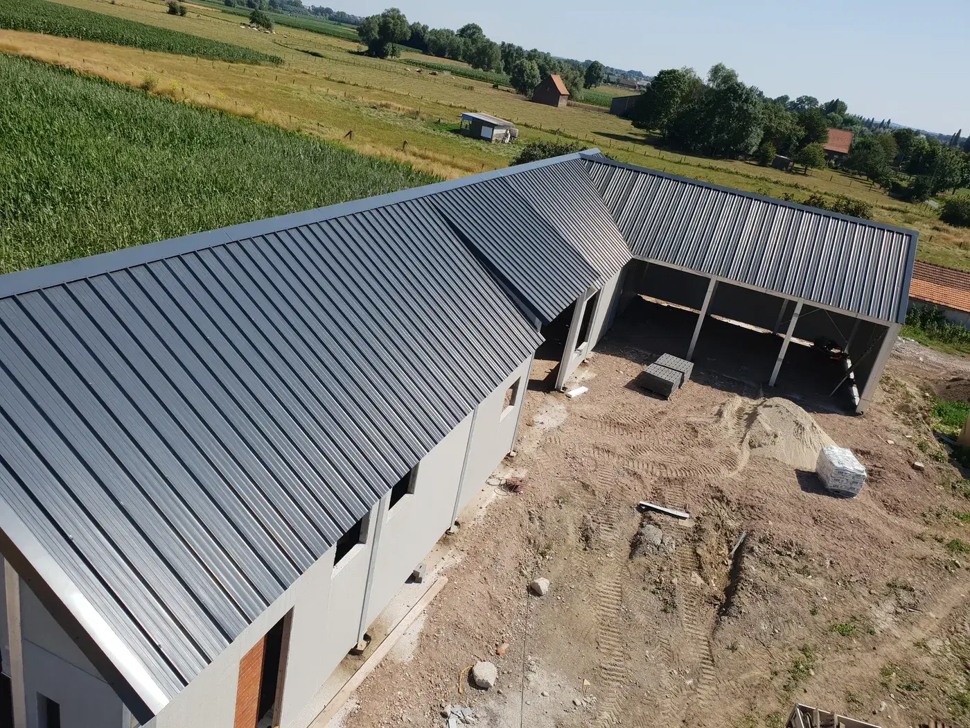 Luchtfoto van een L-vormige hangaar met strak antraciet staaldak in landelijke omgeving