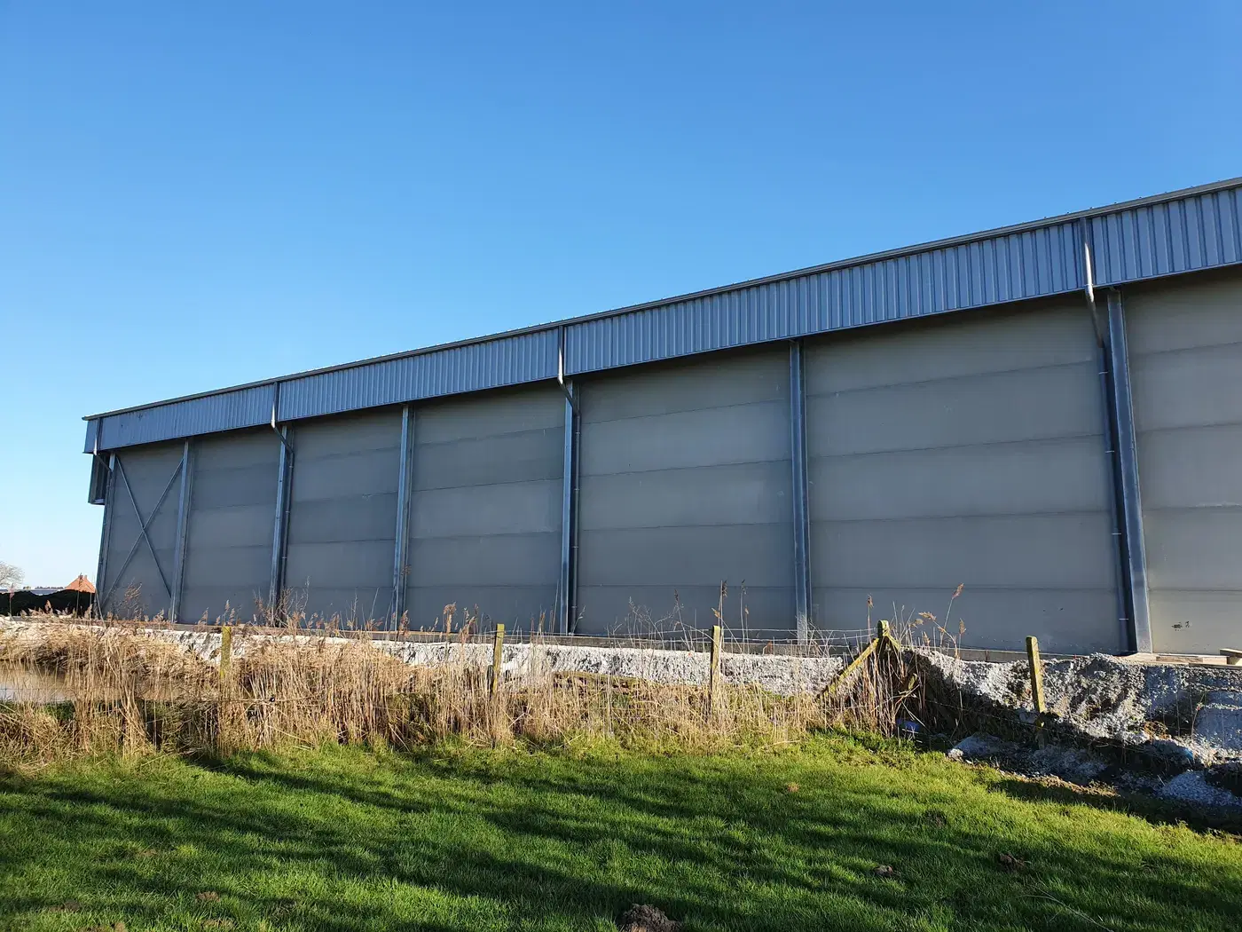 Grote afgewerkte hangaar met betonpanelen en stalen windverbanden onder een blauwe hemel